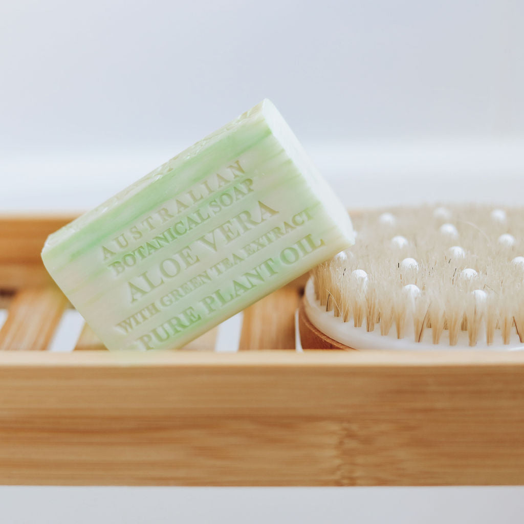 Bar of aloe vera soap on a wooden soap dish with a brush in the background.