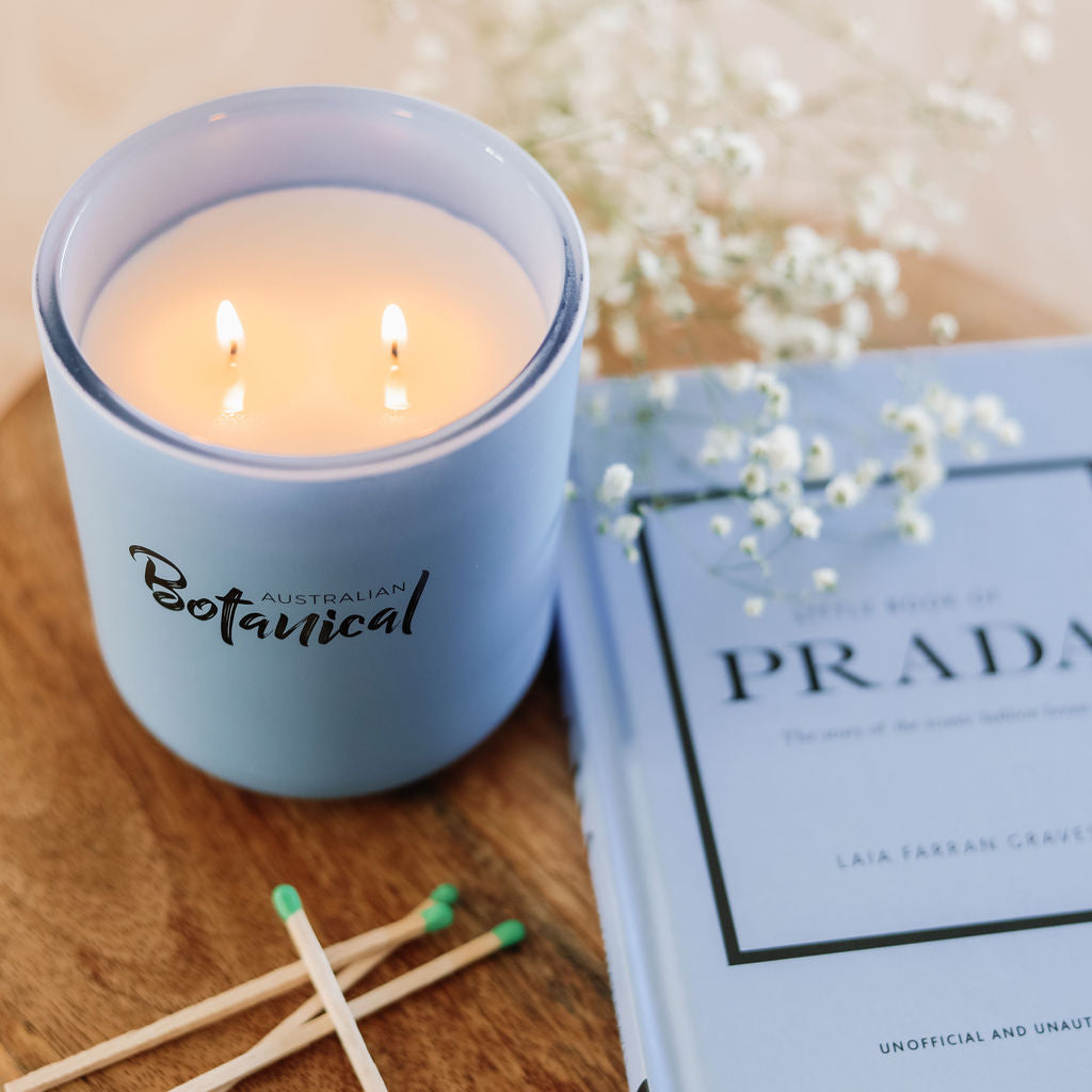 Lit candle in a blue container labeled 'Australian Botanical' next to a book titled 'Prada' on a wooden surface.