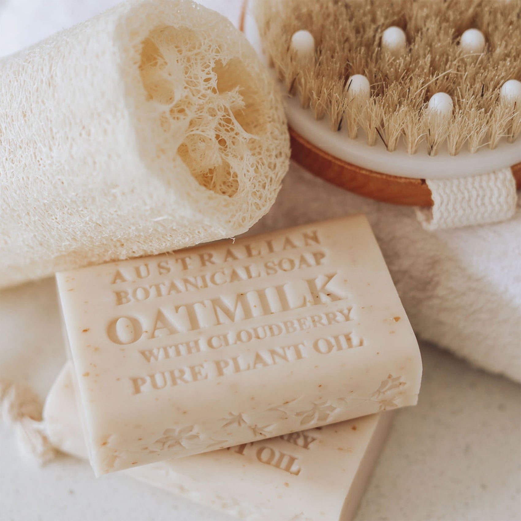 Bar of Australian Botanical Soap with Oatmilk, Cloudberry, and Pure Plant Oil, alongside a loofah and brush on a soft background.