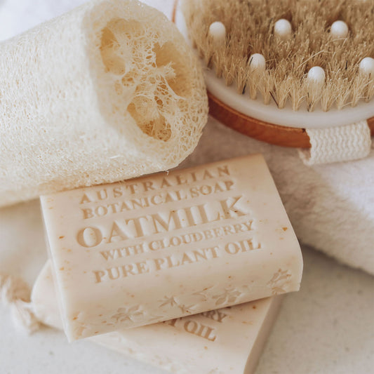 Bar of Australian Botanical Soap with Oatmilk, Cloudberry, and Pure Plant Oil, alongside a loofah and brush on a soft background.