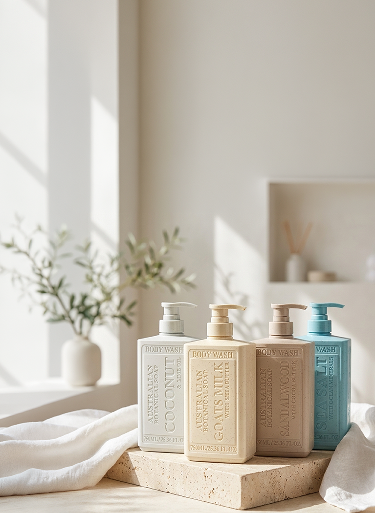 Four pump bottles of body wash in neutral and blue tones arranged on a stone slab in a bright, sunlit bathroom setting with white towels and a plant in the background.