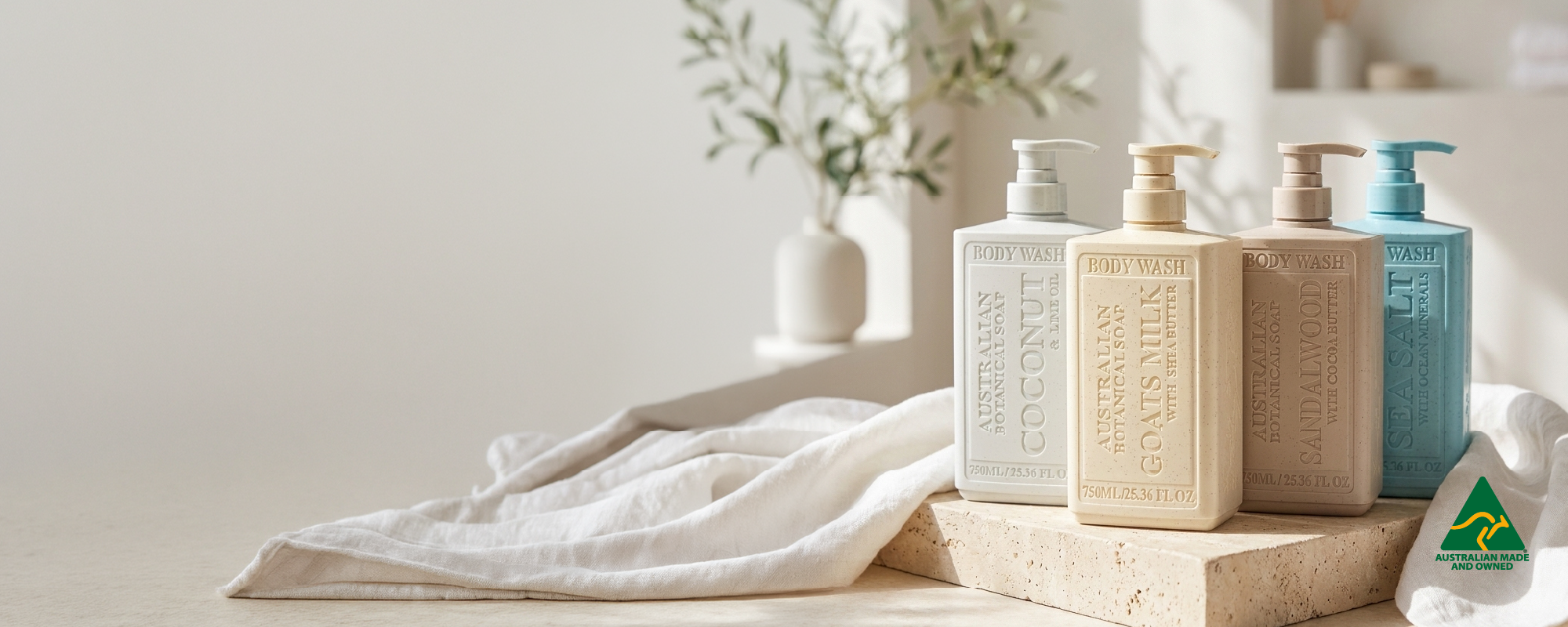 Four textured rectangular pump bottles of soap and body wash in neutral and blue tones, arranged on a stone slab with a white towel and a small plant in the background.