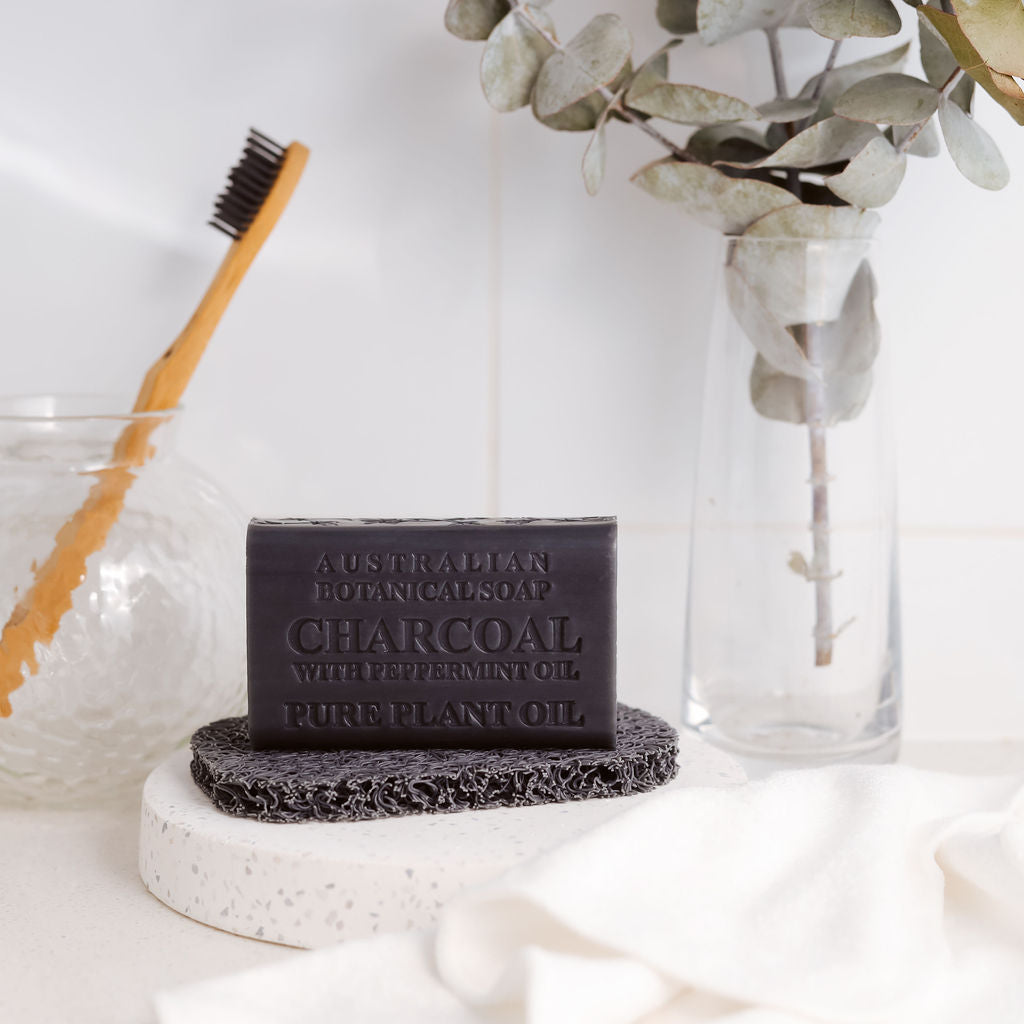 Bar of charcoal soap on a white surface with a toothbrush and plant in the background