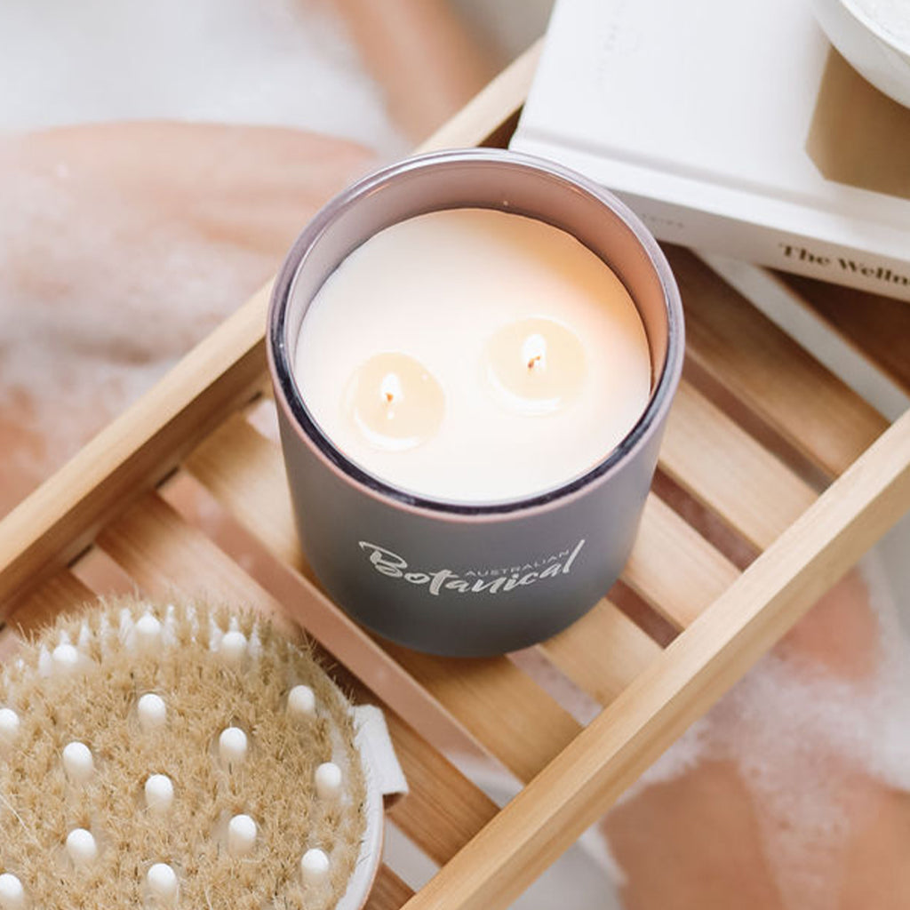 Candle in a dark container labeled 'Botanical' on a wooden tray with a brush and towel in the background.