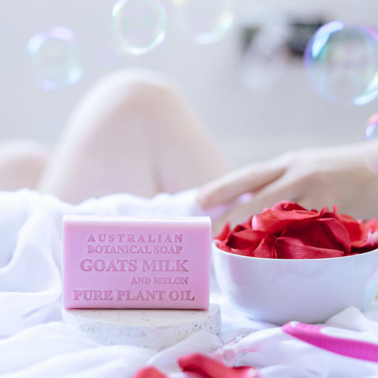 Pink bar of Australian Botanical Soap with Goats Milk and Melon on a white surface with red flowers and bubbles.