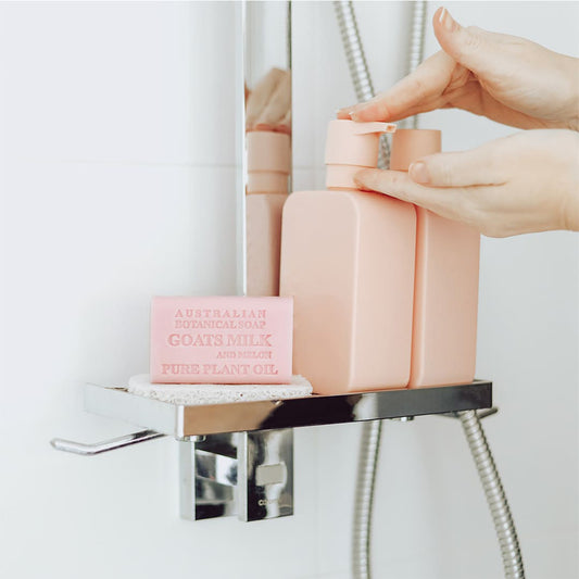 Pink Goats Milk and Melon Bar on a shower shelf with hands pumping pink bottles behind it.