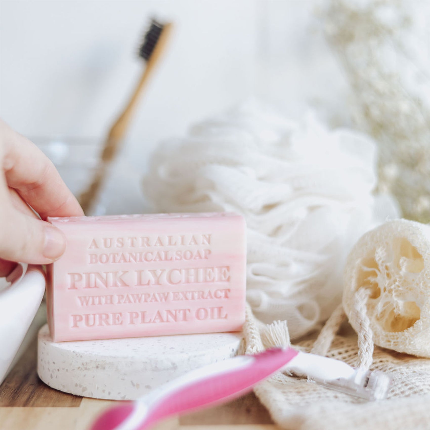Pink soap bar labeled 'Australian Botanical Soap Pink Lychee' held by a hand with a loofah and brush in the background.