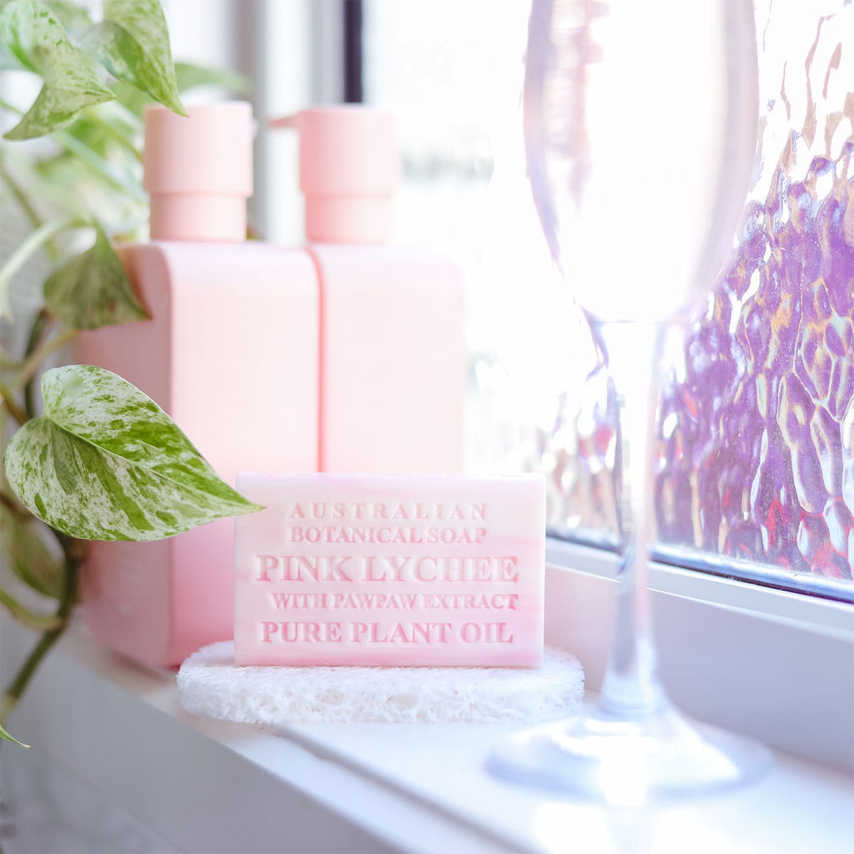 Pink soap bar with packaging on a windowsill with plants and a vase.