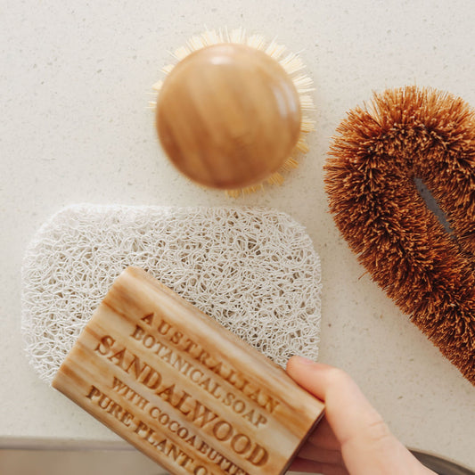 Hand holding a bar of Australian Sandalwood Botanical Soap with Cocoa Butter, surrounded by natural cleaning tools on a light background.