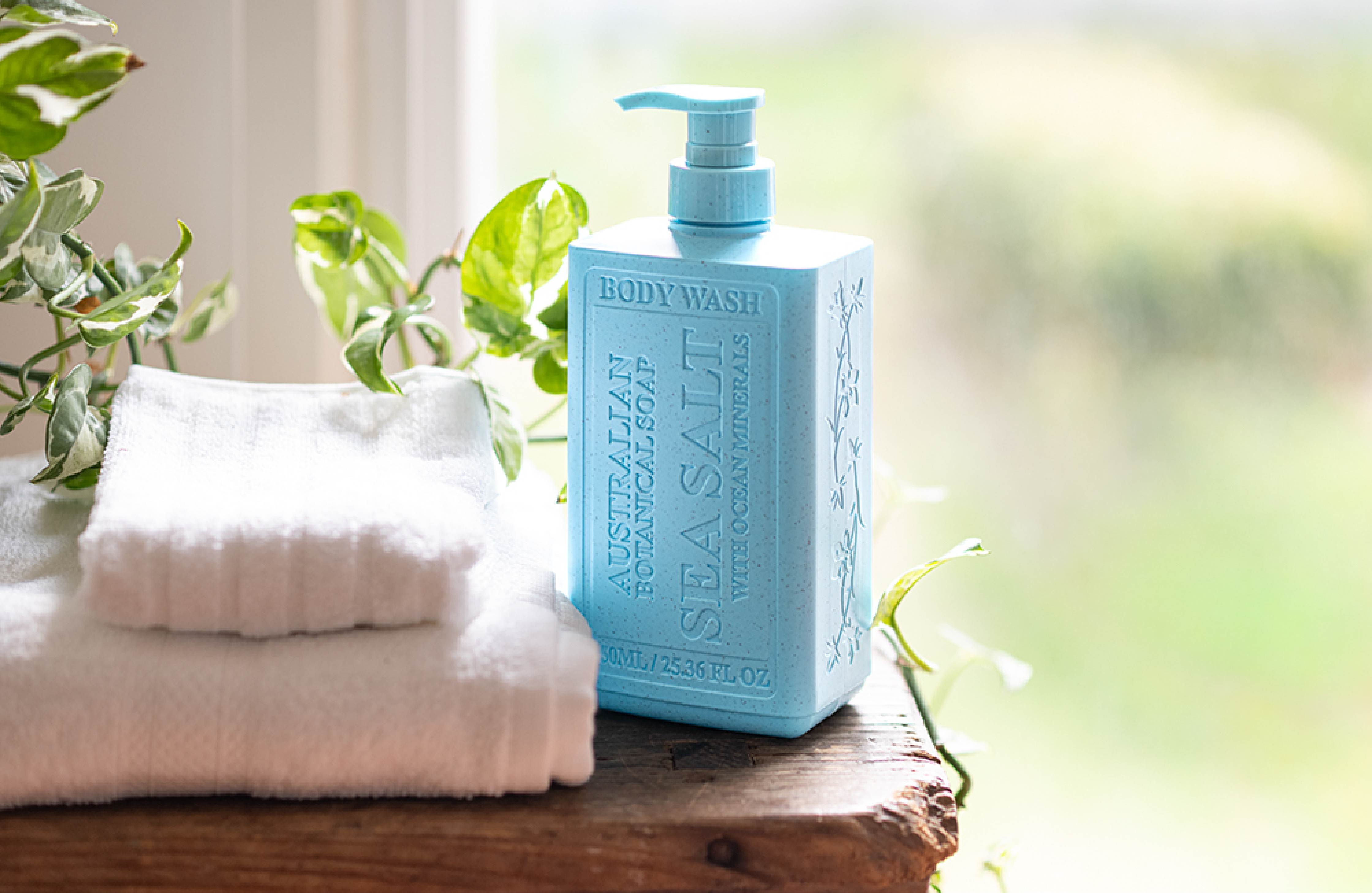 Blue bottle of body wash on a wooden surface with a white towel and green plants in the background