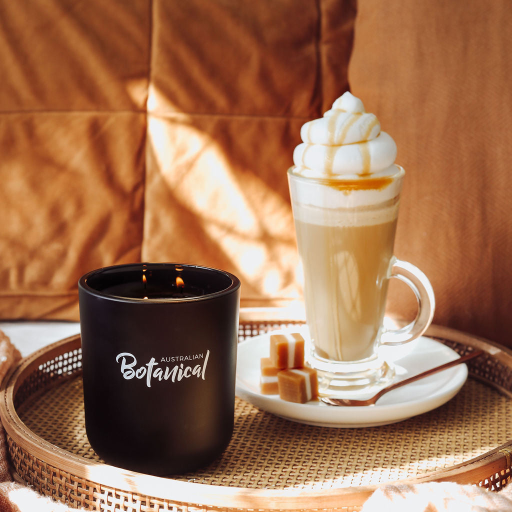 Black candle labeled 'Australian Botanical' next to a glass of latte with whipped cream on a woven tray.
