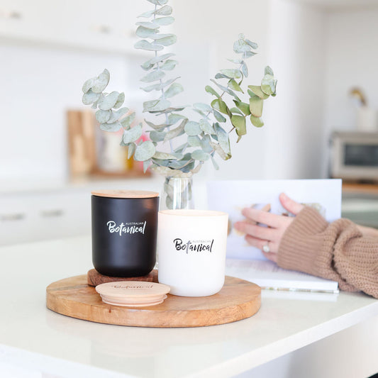Two candles labeled 'Botanical' on a wooden tray with a blurred background of a person and plants.