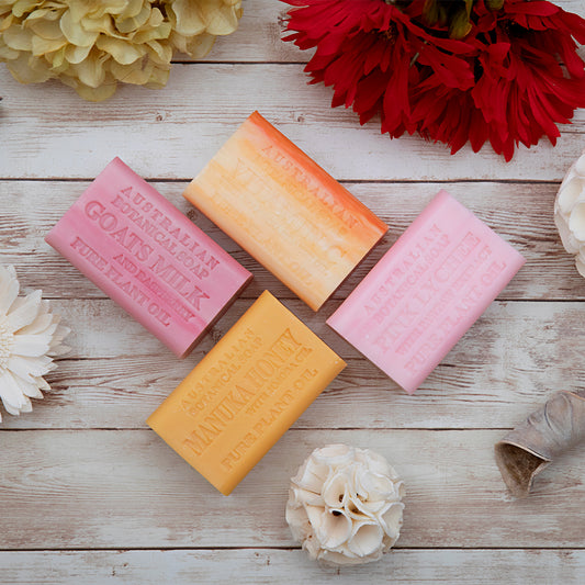 Four bars of soap in pink, orange, and yellow on a wooden surface with flowers around.